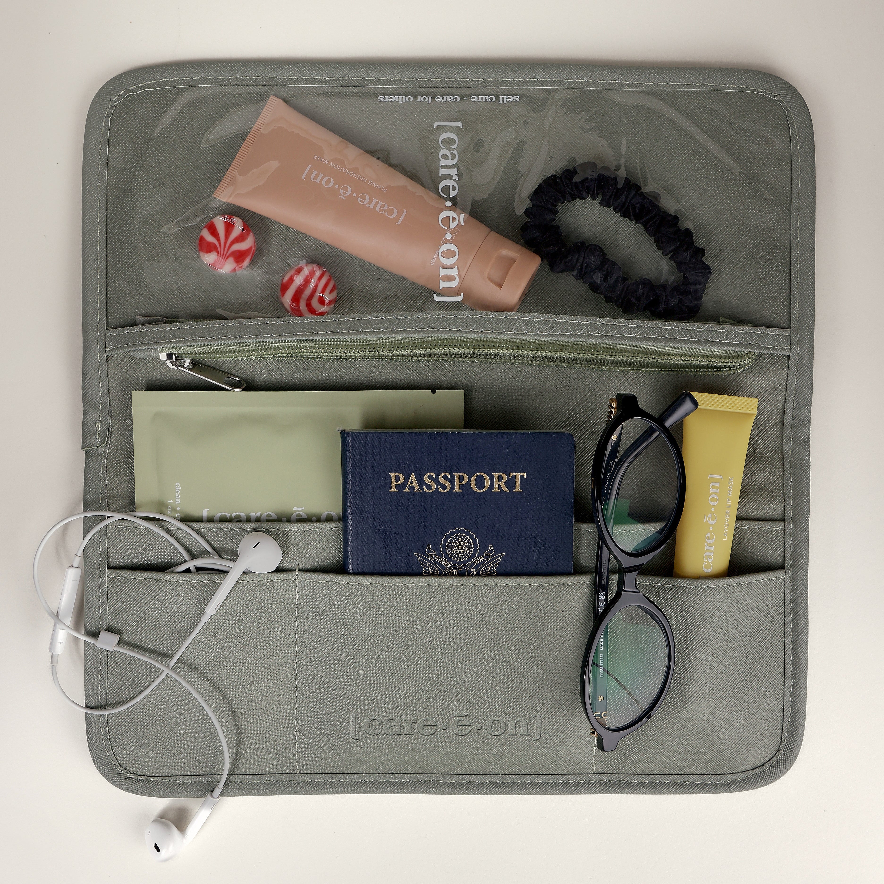 An open gray organizer pouch laid flat on a light beige surface, filled with travel items including a passport, sunglasses, white earbuds, a black scrunchie, a pink tube, and small red-and-white candies. The pouch has multiple zip compartments and pockets.